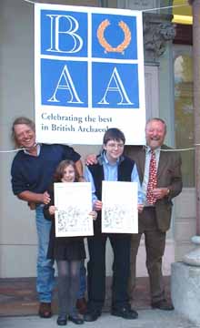 Shows a photo of a girl and a boy holding certificates, in between two men. All are smiling at the camera, with a poster above them saying 'BAA, Celebrating the Best In British Archaeology'.