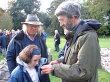 Shows a photo of Beth looking at something an archaeologists is holding, with Phil Harding behind them. They're standing outside, with a crowd in the background.