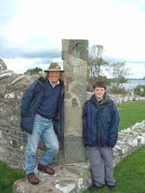 Shows a photo of Christopher and Phil Harding smiling and leaning either side of a standing stone, mounted on a low stone wall.