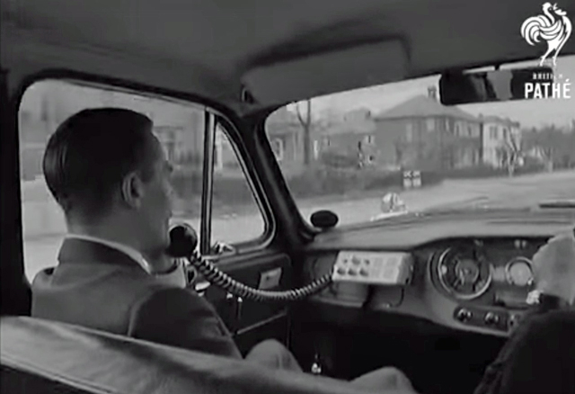 A black and white screenshot showing a man in the passenger side of a car on a telephone