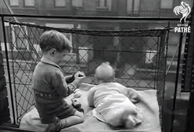 A black and whote screen shot showing two small children in a cage hung out a window. 