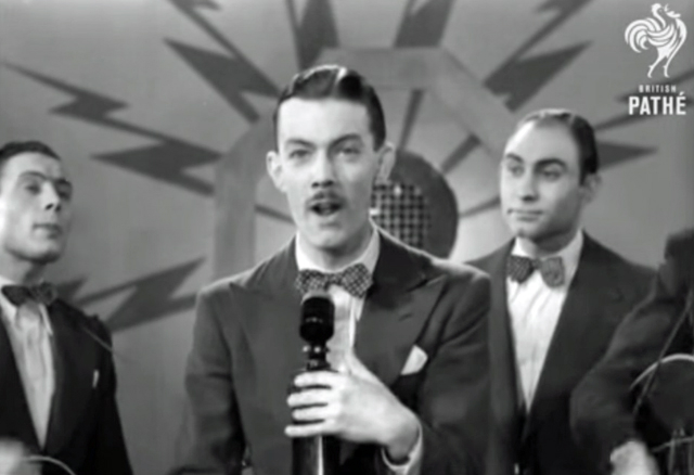 A black and white screenshot showing a man standing with a microphone