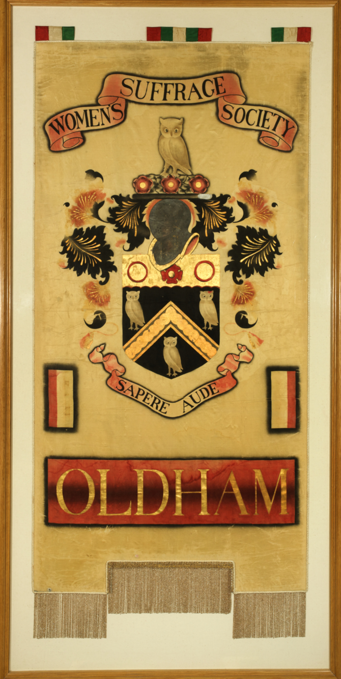 A gold banner created in 1913 by Oldham Womens Suffrage Society. The banner is tall and rectangular decorated with the Oldham crest and the latin phrase 'Sapere Aude' meaning 'Dare to Know'.