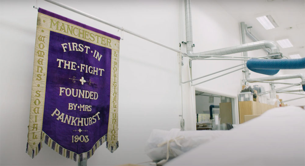 a purple banner on a white brick wall reading "FIRST IN THE FIGHT FOUNDED BY MRS PANKHURST 1903" in gold lettering